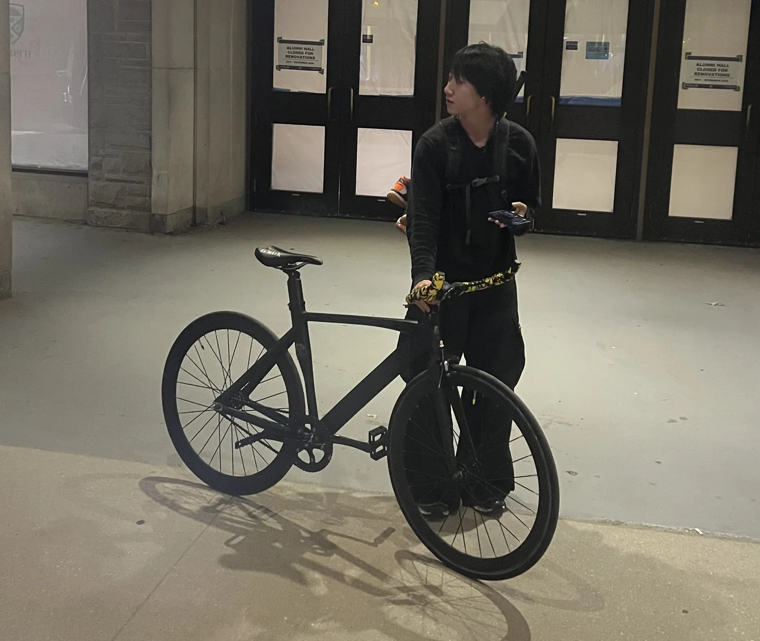 David standing with his matte black track bike at night, outside Alumni Hall at Western